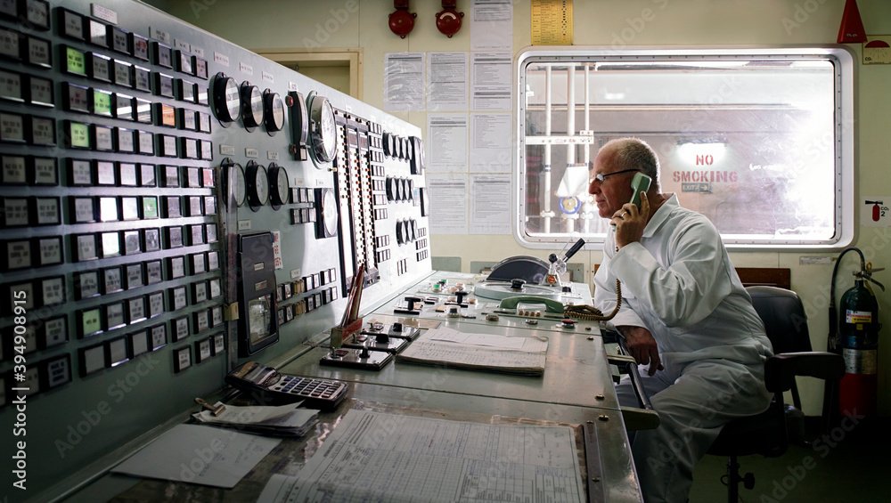 Senior engineer taking phone instructions whilst monitoring controls in ship's engine room.